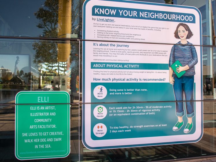 Image of A large sign on a glass wall displays health tips about physical activity, with an illustrated woman holding a book. A smaller sign introduces Elli as an artist and community arts facilitator who loves being creative, walking her dog, and swimming.