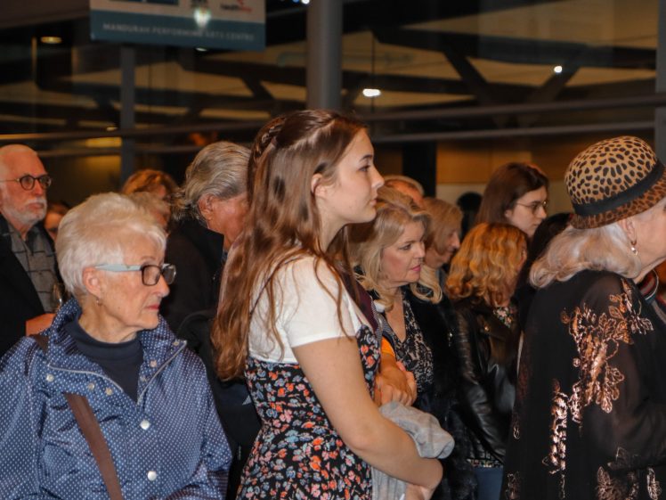 Image of A diverse group of people, both young and elderly, stand closely together indoors, attentively facing the same direction. The scene includes women in patterned clothing and jackets, with warm lighting in the background.