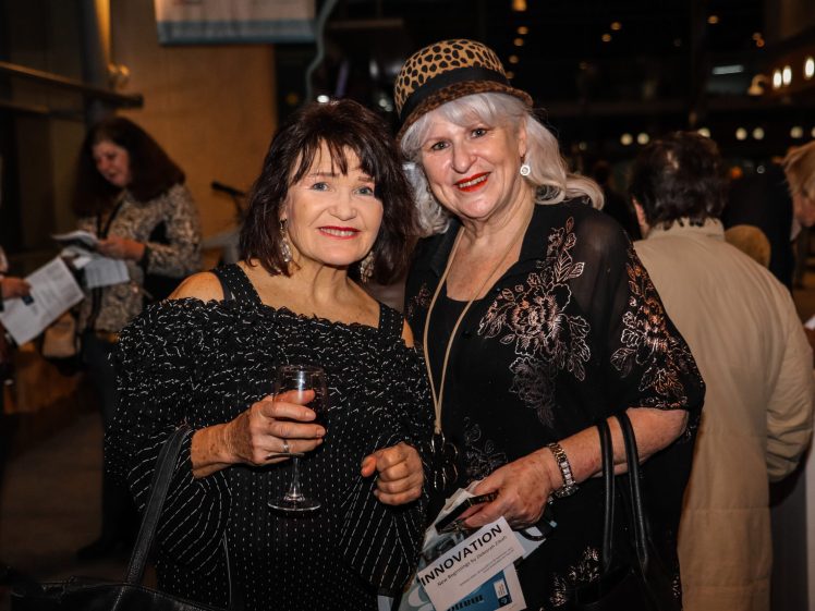 Image of Two older women dressed in black and gold pose and smile at an indoor event. One holds a glass of wine, the other wears a patterned hat. People in the background are mingling and holding programmes.