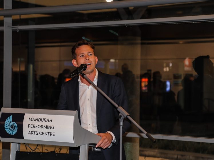 Image of A man in a suit speaks into a microphone at a lectern labelled Mandurah Performing Arts Centre inside a modern building with glass walls.
