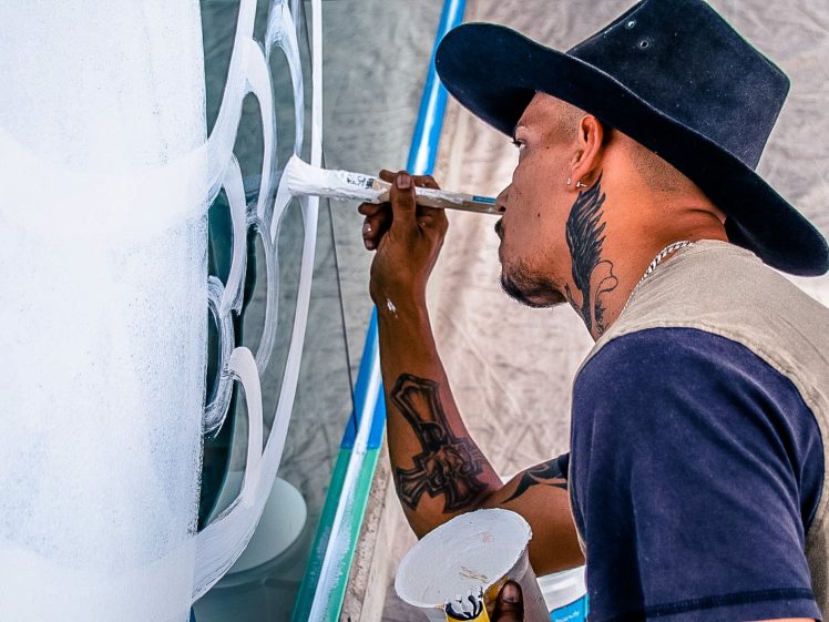 Image of A man wearing a black hat paints a mural with white paint. He holds a brush in one hand and a paint palette in the other. Tattoos are visible on his neck and arm.