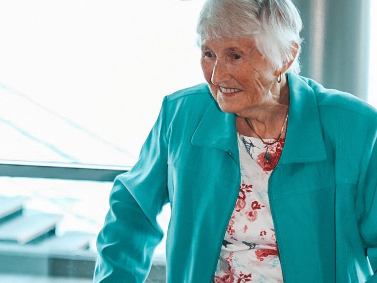 Image of An elderly woman with short white hair, wearing a turquoise jacket over a floral top, smiles while holding onto a Zimmer frame. She stands indoors near a large window with a bright, blurred background.