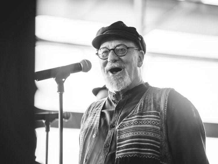 Image of An older man with a grey beard, glasses, and a cap sings into a microphone. He wears a patterned waistcoat and shirt, and appears joyful. The image is black and white with a blurred background.