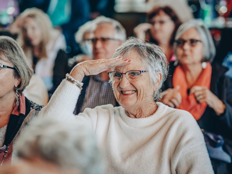 Image of An older woman in a white jumper smiles and shades her eyes with her hand while sitting among a group of people at an indoor event.