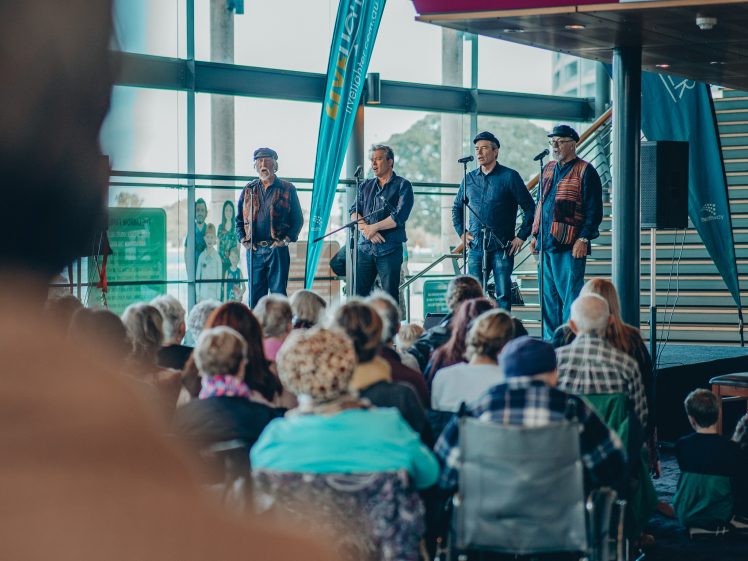 Image of Four men stand and sing on a stage indoors, facing an audience seated in rows. The venue has large windows, banners, and a staircase. The audience includes people of various ages, some using wheelchairs.