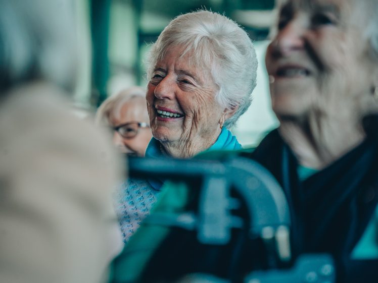 Image of A group of elderly women sitting together, smiling and laughing, with one woman in focus wearing a light blue scarf and white hair. The atmosphere appears joyful and friendly.