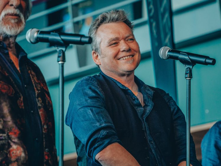 Image of A man with short, greyish hair smiles whilst standing at a microphone. He is wearing a dark shirt and waistcoat, and appears to be speaking or singing indoors, with another person partially visible beside him.