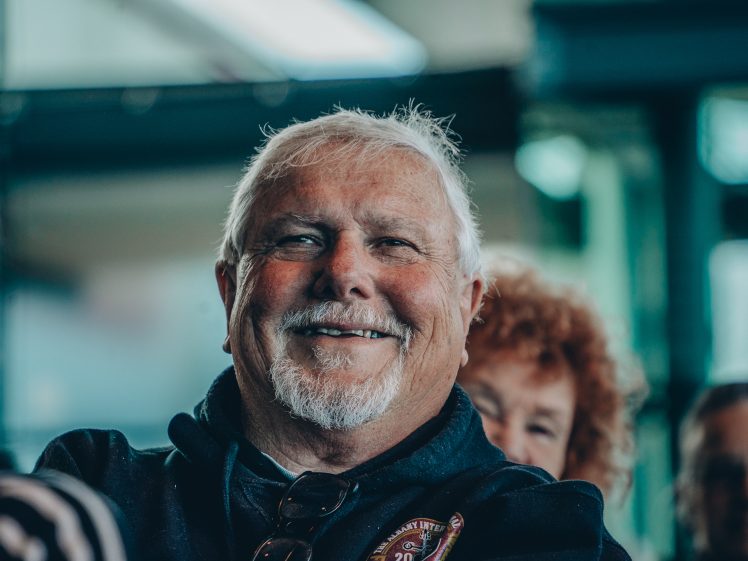 Image of An older man with white hair and a beard smiles warmly at the camera, wearing a dark hoodie and sunglasses. He is indoors, with other people and blurred windows in the background.
