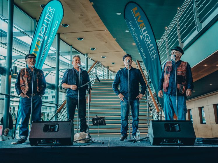 Image of Four men stand singing on a stage with microphones in front of a staircase. Two blue LiveLighter banners are displayed behind them, and large windows brighten the modern indoor space.