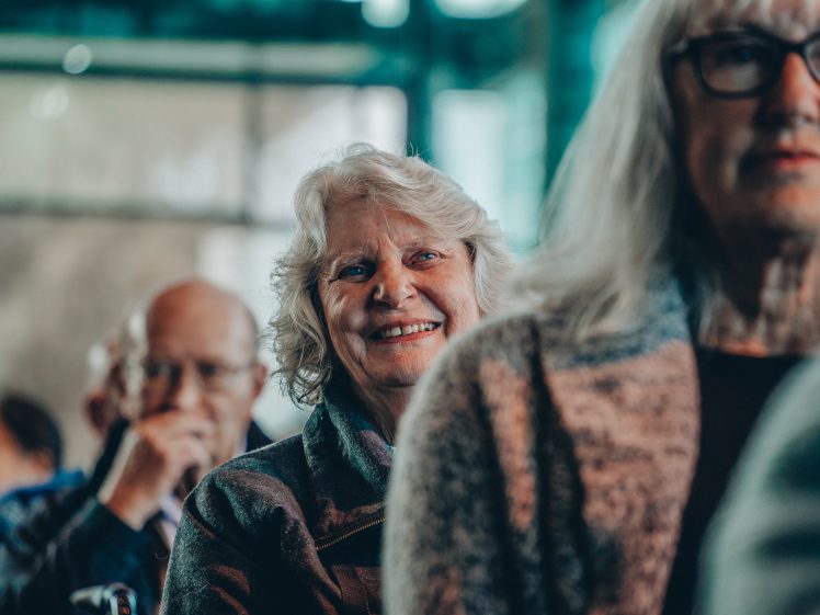 Image of An older woman with white hair sits among a group of people, smiling warmly. The focus is on her, while others around her, including a man and another woman, are blurred in the foreground and background.