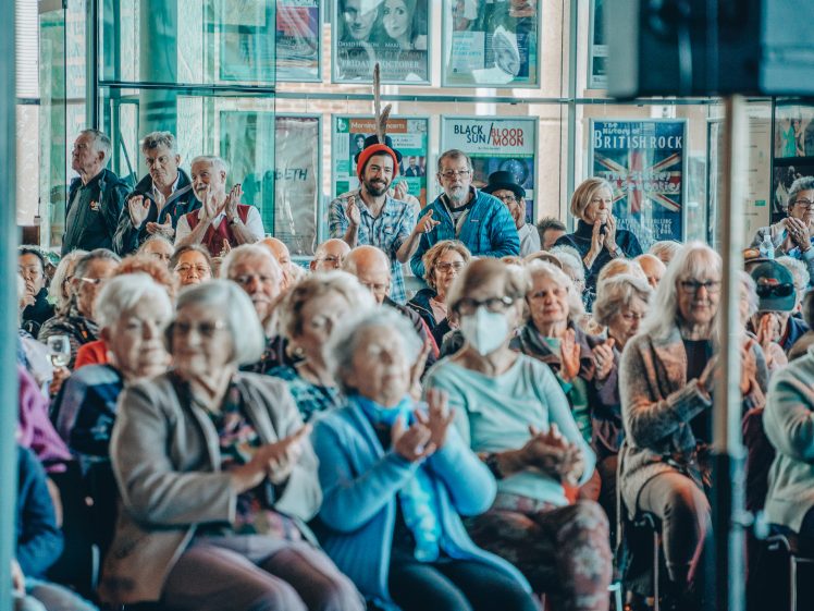 Image of A large group of elderly people, some wearing masks, are seated indoors and applauding. Posters and windows are visible in the background, creating a lively and engaged atmosphere at the event.