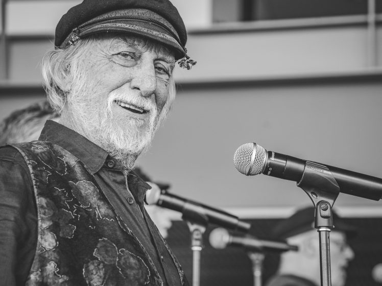 Image of An older man with a white beard and a hat smiles while standing at a microphone. Two other people, also wearing hats, are blurred in the background. The image is in black and white.