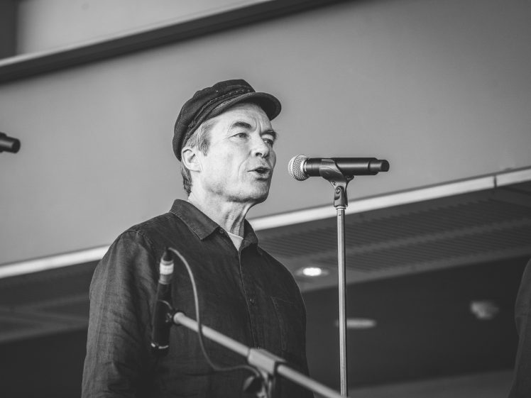 Image of A man wearing a cap and button-up shirt sings or speaks into a microphone on a stage, with another microphone stand visible nearby; the image is in black and white.