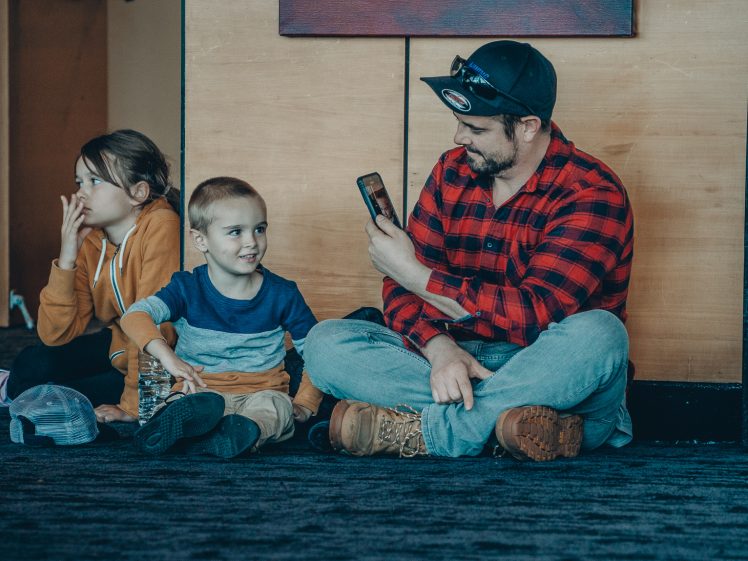 Image of A man in a red checked shirt sits on the floor with two children, smiling and holding up a mobile, possibly taking a photo or video. The children sit beside him, one looking at the camera, the other gazing to the side.