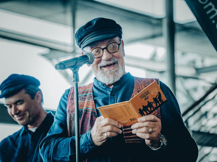 Image of An older man with a white beard and glasses, wearing a dark cap and patterned waistcoat, reads from a booklet into a microphone. Another smiling man in a similar outfit stands beside him. They appear to be performing indoors.