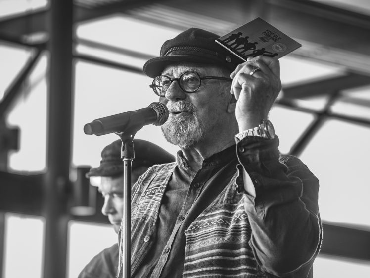 Image of A bearded man wearing glasses, a hat, and a patterned shirt speaks into a microphone on stage while holding up a CD case. The image is in black and white. Another person is blurred in the background.