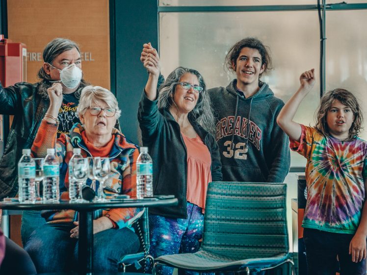 Image of A group of five people, some standing and some seated, cheer with raised hands. They look happy and engaged. Bottles of water are on a table in front of them. One person wears a mask, another has a tie-dye shirt.