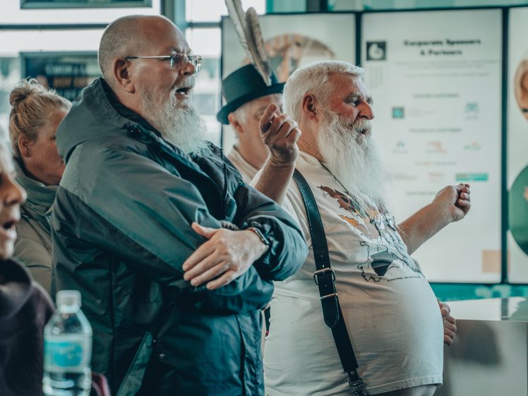 Image of A group of older adults, including two men with long white beards, stand indoors, engaged in conversation or watching something intently. One man wears a jacket; the other wears a T-shirt with braces.