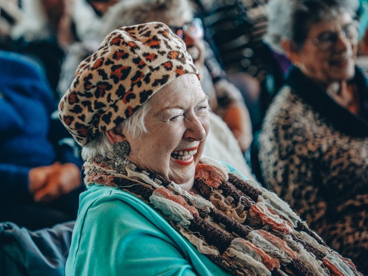 Image of An elderly woman wearing a leopard-print hat and colourful scarf smiles and laughs whilst sitting in a crowd, surrounded by other older adults.