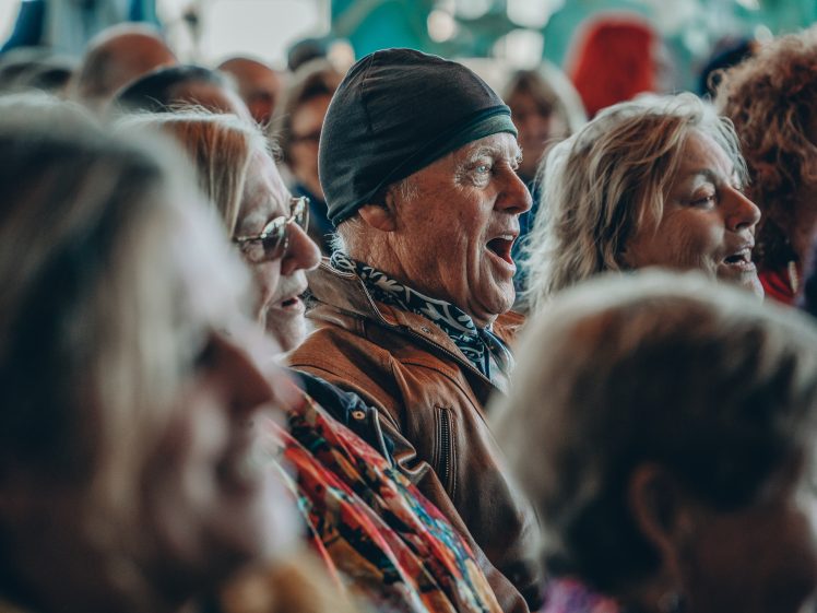 Image of A group of older adults sitting closely together, some smiling and one man in a beanie and brown jacket singing or speaking enthusiastically in the centre, all appearing engaged in an event or gathering.