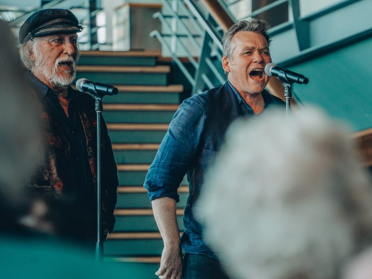Image of Two men stand side by side singing into microphones in front of a staircase, with blurred audience members in the foreground.