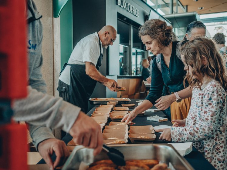 Image of A woman and a young girl select food from trays at a serving table, while volunteers serve meals in a bright indoor setting near a box office. Other people serve and queue.