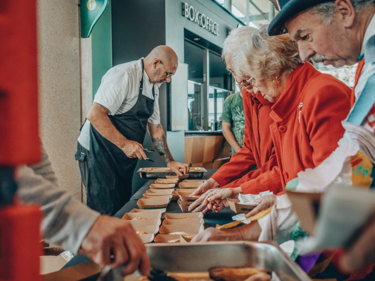Image of A group of people, including elderly women in red coats, stand at a food counter serving and preparing meals in cardboard trays. A chef in an apron slices food behind the counter near a box office.