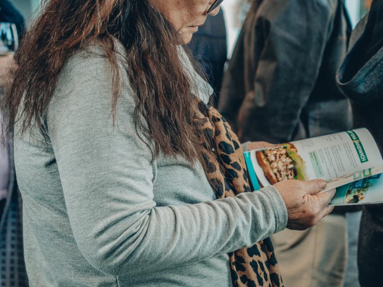 Image of A woman with long brown hair and glasses, wearing a grey jumper and holding a leopard print scarf, looks down at an open leaflet while standing among other people indoors.
