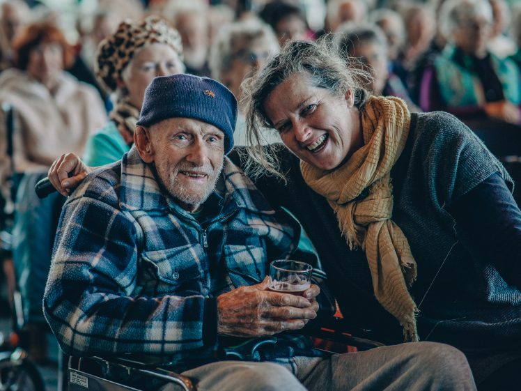 Image of An elderly man in a wheelchair, wearing a blue checked jacket and blue hat, smiles whilst holding a drink. A woman beside him, wearing a yellow scarf, leans in and smiles. A group of people is visible in the background.