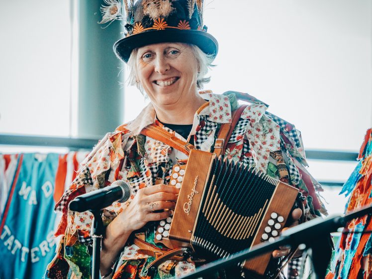 Image of A smiling person in colourful patchwork clothing and a decorated hat plays an accordion in front of a microphone, with bright banners in the background.