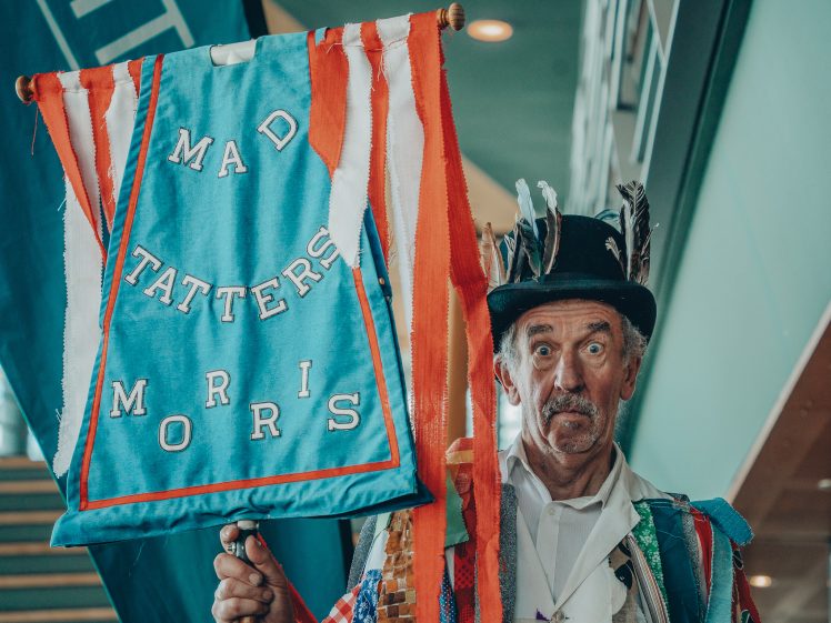 Image of An older man in a decorated costume with feathers in his hat holds a blue banner reading MAD TATTERS MORRIS. He stands indoors, looking at the camera. The banner is trimmed with red, white, and orange ribbons.