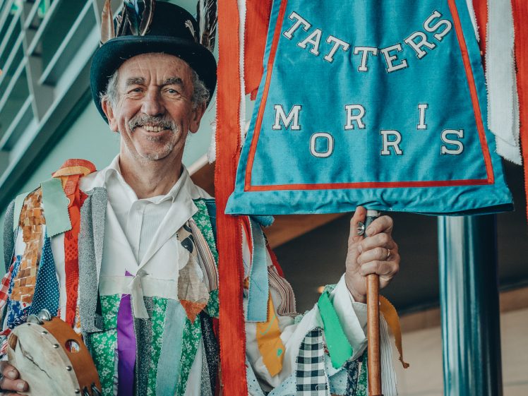 Image of An older man in a colourful patchwork costume and feathered hat holds a tambourine and a banner reading MAD TATTERS MORRIS, smiling indoors near a staircase.