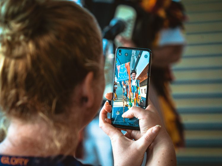 Image of A person with light brown hair holds up a mobile phone, taking a photo of two people standing on stairs; the phone screen clearly displays the posing pair in colourful outfits.