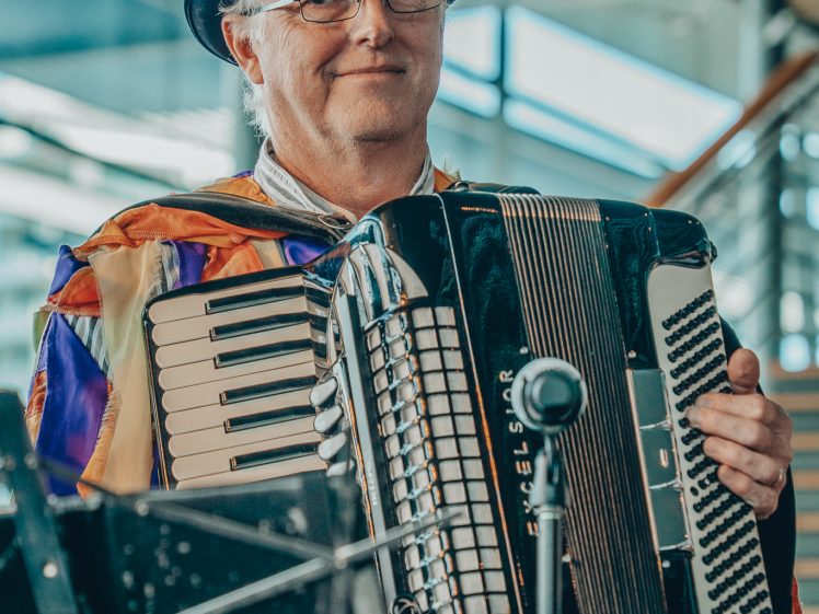 Image of A man wearing a colourful costume, glasses, and a black hat with a feather smiles whilst playing an accordion indoors. There is a microphone and a music stand in front of him, with stairs and windows in the background.