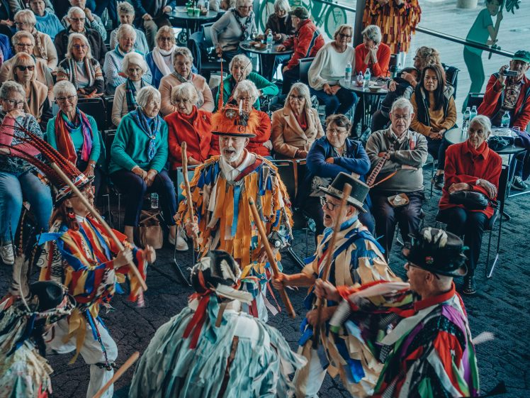Image of A group of people in colourful, ribboned costumes perform a traditional dance with sticks for an audience of older adults who are seated and watching attentively in a bright indoor setting.