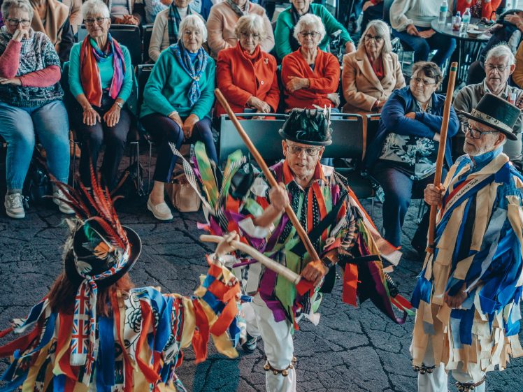 Image of Three dancers in colourful, traditional costumes perform with sticks in front of an audience of elderly people, most wearing bright jumpers, seated and watching intently. The performers' outfits feature ribbons and patterns.