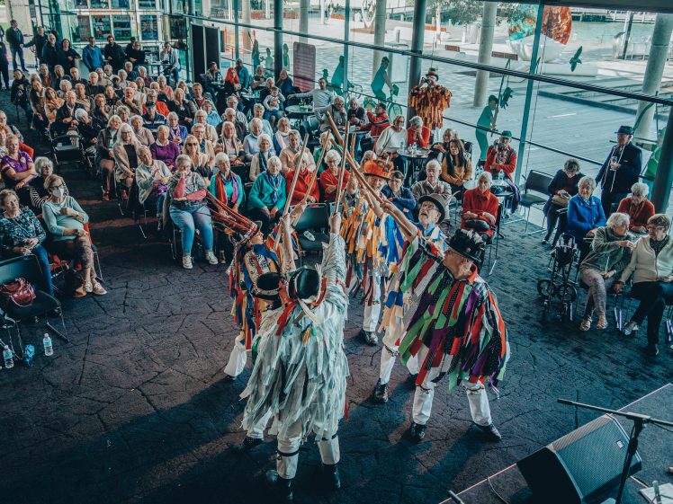 Image of A group of dancers in colourful, ribboned costumes perform in front of an audience seated indoors, with large windows revealing an outdoor scene. The audience watches attentively, some seated in wheelchairs.