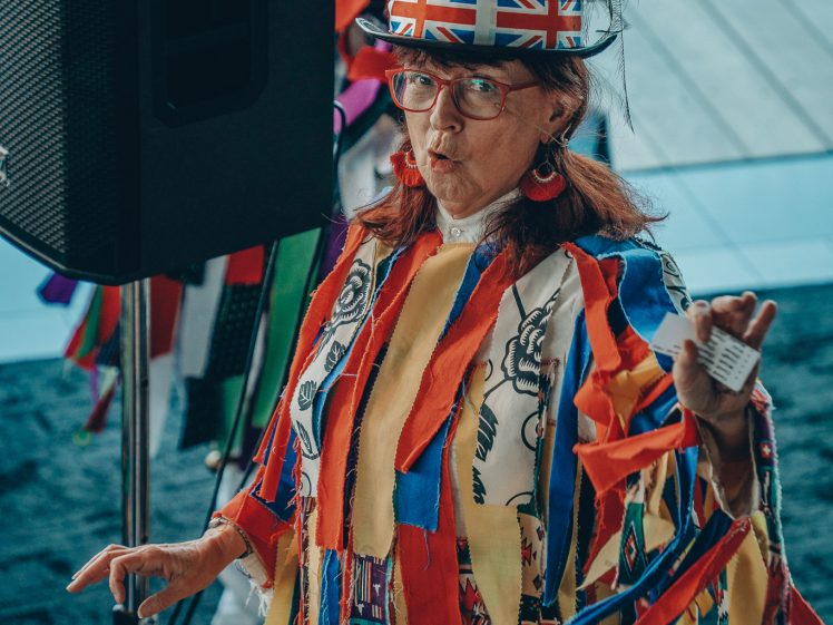 Image of A person in a colourful, fringed outfit and a headdress with red feathers and a Union Jack band stands indoors, holding a small item, with expressive facial features and spectacles.