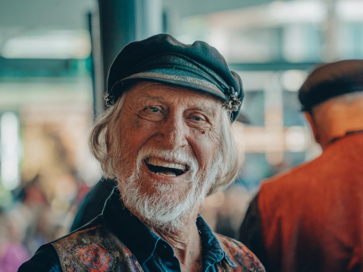 Image of An older man with a white beard and hair, wearing a black cap and patterned waistcoat, smiles warmly at the camera in a brightly lit indoor setting.
