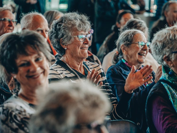 Image of A group of older adults, mostly women, sit close together indoors, smiling and clapping whilst enjoying an event. They wear glasses and casual clothing, and the atmosphere appears joyful and lively.