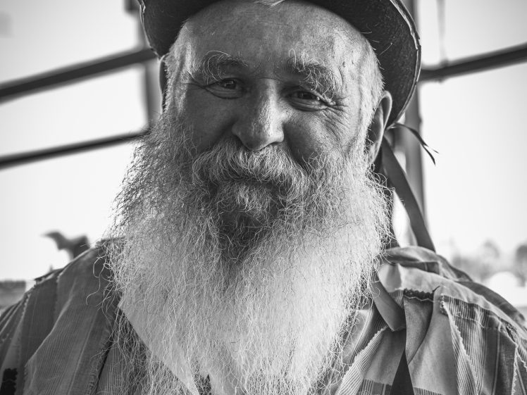 Image of An older man with a long white beard, wearing a hat decorated with feathers and a striped shirt, smiles warmly at the camera. The image is in black and white with a blurred background.