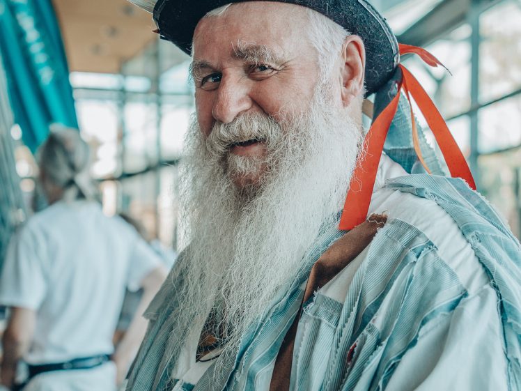 Image of An older man with a long white beard wears a decorated hat with feathers and ribbons, and a costume with tattered fabric strips. He smiles while standing indoors at what appears to be a festive or cultural event.