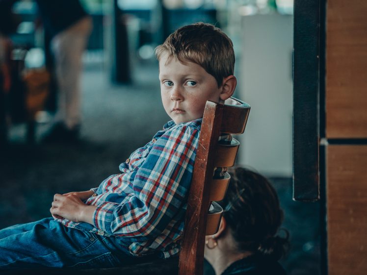 Image of A young boy in a checked shirt sits sideways on a chair, gazing towards the camera with a serious expression. Another person, mostly out of frame, sits nearby with their head slightly lowered.
