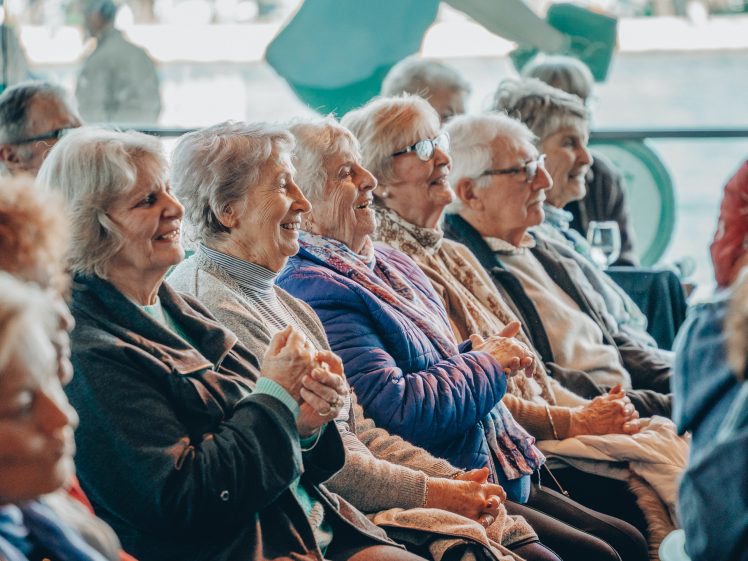 Image of A group of elderly people sitting closely together, smiling and laughing as they watch something off camera in a bright, indoor setting.
