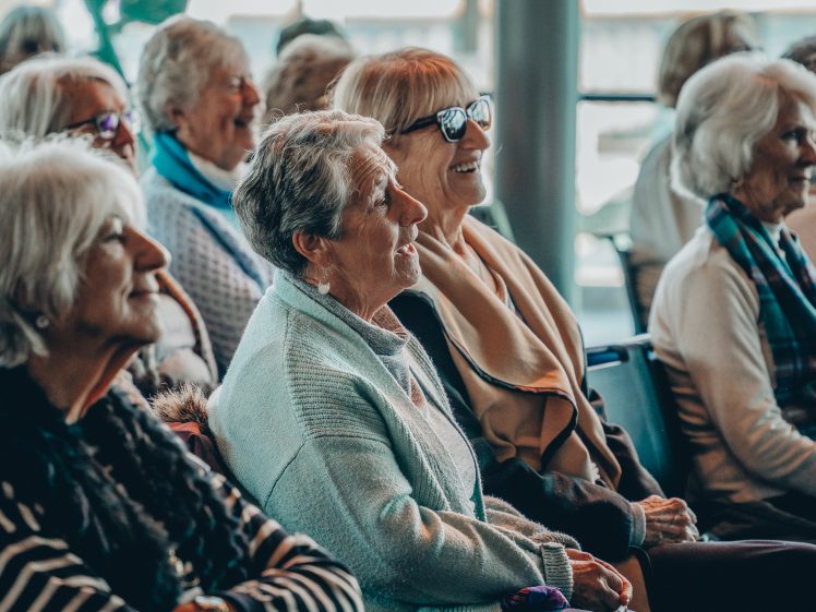 Image of A group of older women sitting together, smiling and laughing, as they watch an event or presentation in a bright, indoor setting.