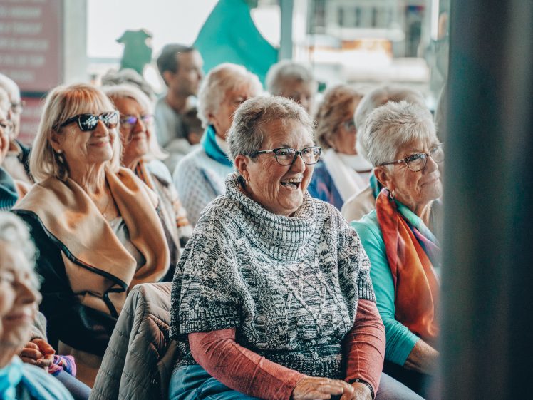 Image of A group of older women sit together indoors, smiling and laughing. They wear glasses, casual clothes, and colourful scarves, appearing to enjoy a lively event or gathering.