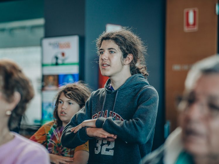 Image of Two young people stand indoors, one in a grey hoodie with arms crossed and another in a colourful shirt behind them. Both appear focused, with other people blurred in the foreground.