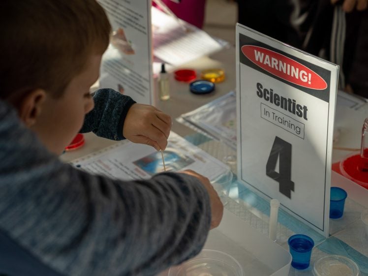 Image of A child wearing a blue jumper uses a dropper at a science activity table labelled Warning! Scientist in Training 4, surrounded by lab materials and colourful containers.