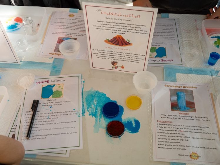 Image of A tabletop set-up for science experiments with instruction sheets, plastic cups, a spoon, coloured powders, and spilt blue liquid, suggesting activities like saltwater eruptions and colour mixing for children.