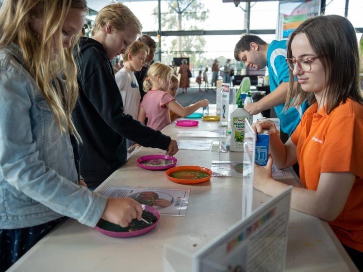 Image of Children stand at a table, engaging in a hands-on science activity with colourful plates. An adult in an orange shirt assists, while another adult in blue helps other children in the background. The setting appears to be a busy indoor event.